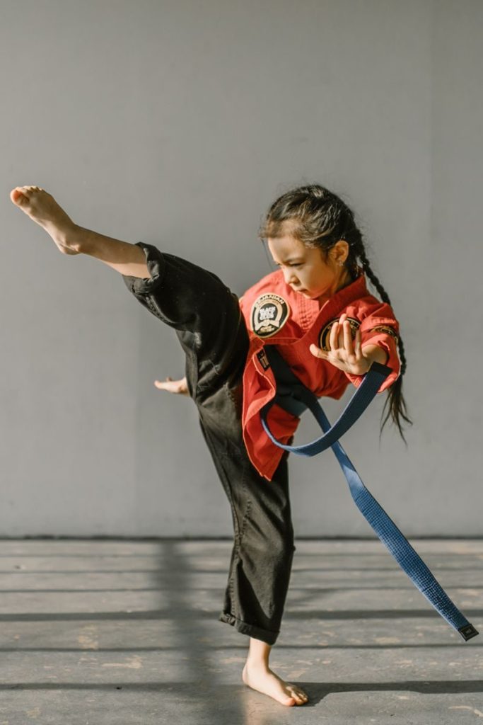 Child practicing a high kick in martial arts dojo wearing a blue belt.